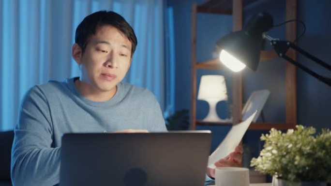 年轻的亚洲商人将在线视频通话会议的任务集中在晚上加班在客厅的笔记本电脑中与同事一起工作。