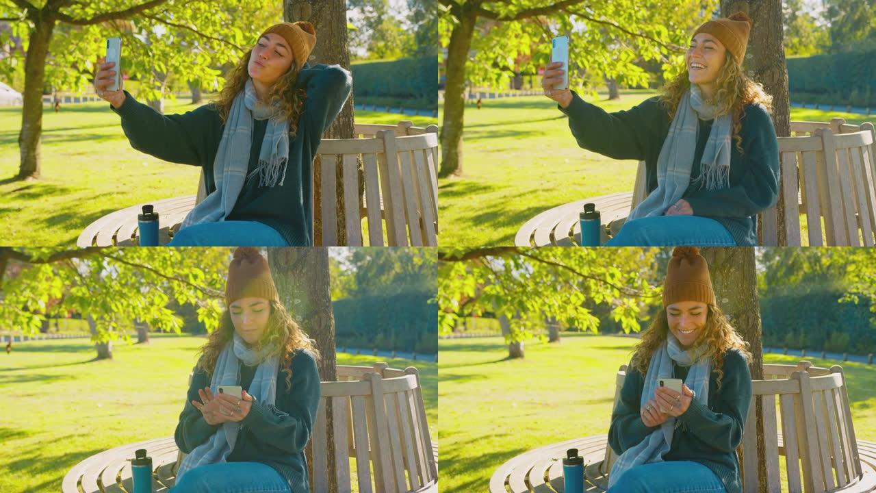 女子坐在秋园的长凳上摆姿势拍照在手机上发布到社交媒体上