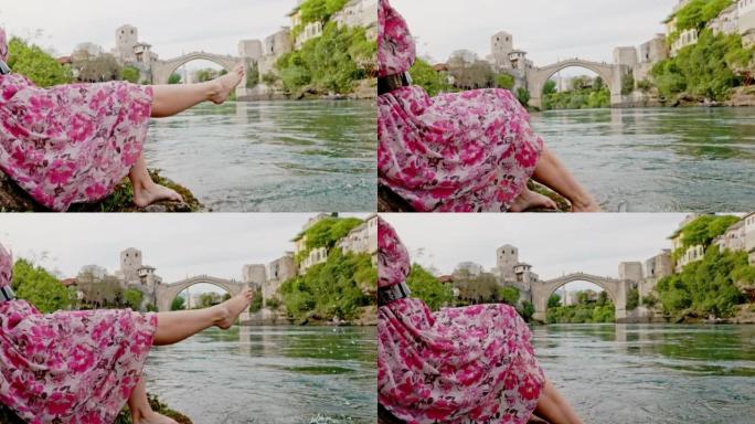 穿着粉红色连衣裙的特写女人坐在neretva河岸，将赤脚伸入水中，将腿向上移动，溅水，莫斯塔尔市在背