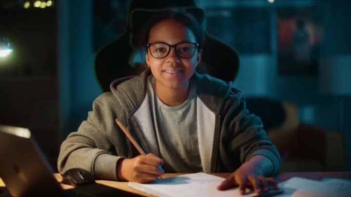 年轻的多种族黑人女孩在家里黑暗舒适的房间里用笔记本电脑用铅笔在笔记本上写下作业。她很高兴，看着相机微