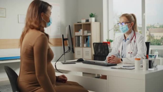 一位女家庭医生正在一家诊所与年轻的怀孕病人交谈。两人都要戴口罩。穿着白大褂的医生坐在医院办公室的电脑