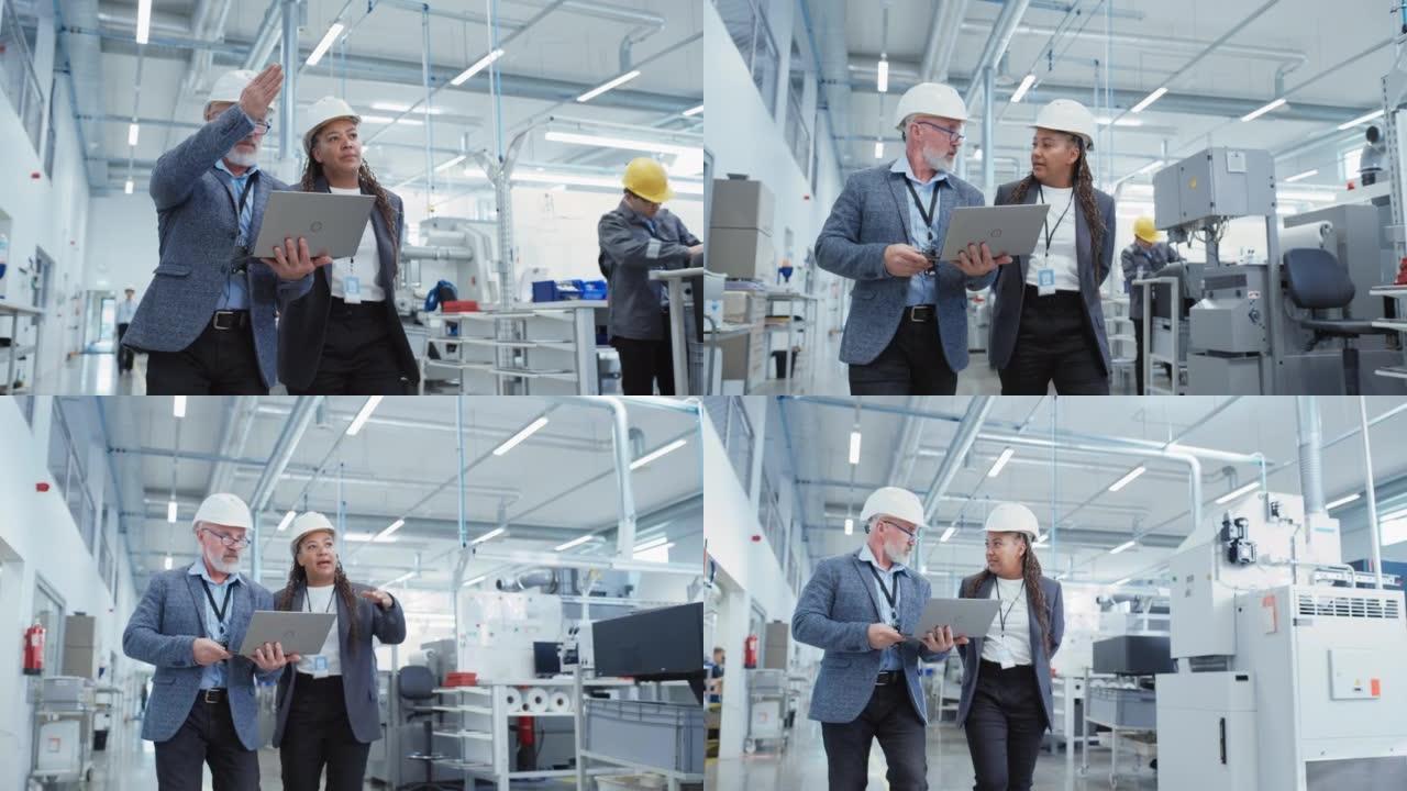 两名戴着安全帽的不同重工业工程师的肖像，他们戴着笔记本电脑走路，在工厂里聊天。两名制造业员工在工作的