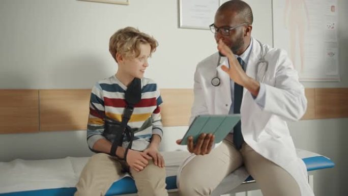 友好的非裔美国家庭医生与一个带手臂支架的小男孩交谈，并在平板电脑上显示测试结果。快乐的医疗保健医生在