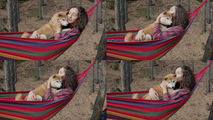 美丽的年轻女士拥抱可爱的柴犬狗狗在秋日在森林里放松的吊床上摇摆