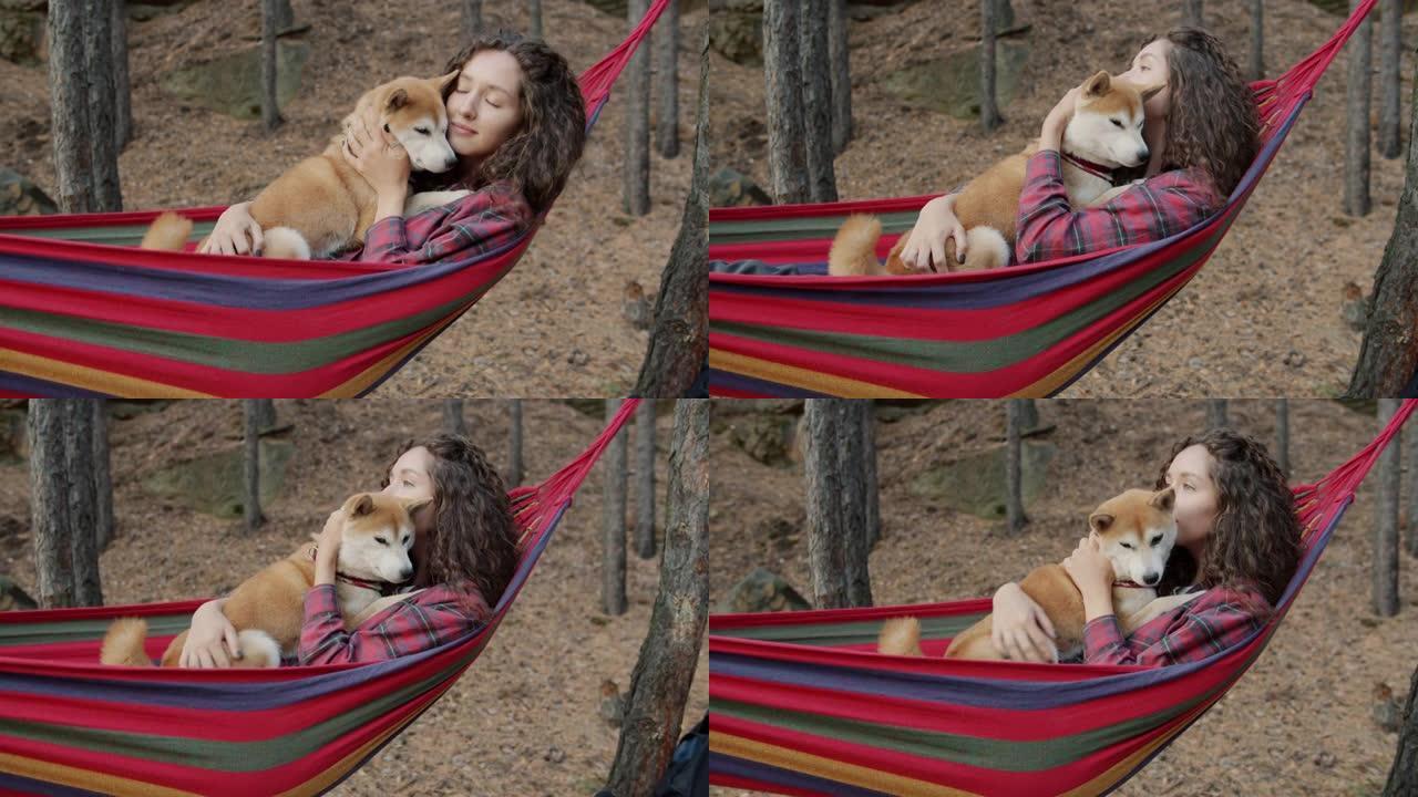 美丽的年轻女士拥抱可爱的柴犬狗狗在秋日在森林里放松的吊床上摇摆