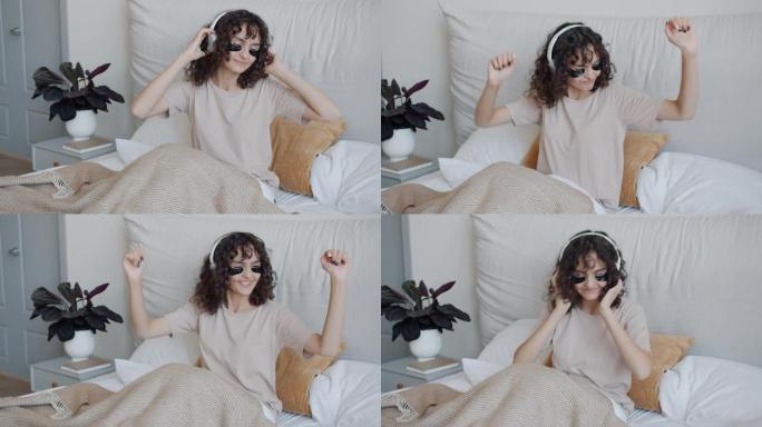 戴着眼罩的快乐女孩通过耳机在床上跳舞听音乐