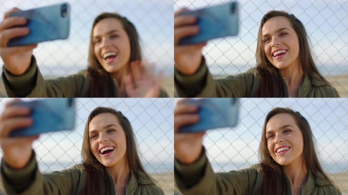 一个快乐的年轻女子用手机打视频电话并挥手致意。一名妇女在智能手机上进行视频通话时挥舞着，站在篱笆旁。
