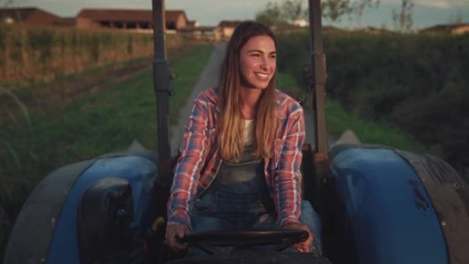 一名年轻女子在绿色的玉米田旁在路上驾驶拖拉机的中等镜头。女农民在清晨开始她的工作任务。一个反对陈规定