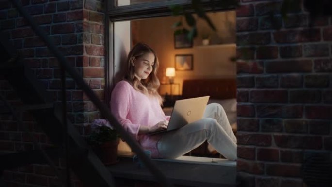 美丽的年轻白人妇女晚上坐在敞开的窗台上使用笔记本电脑。穿着舒适衣服的创意女性微笑着在电子日记中写下日