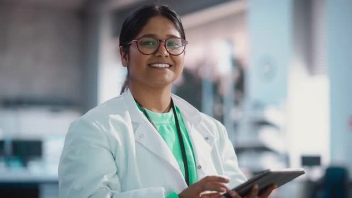 印度年轻女科学家的肖像使用数字平板电脑，看着相机，在实验室里微笑。自信的职业女性担任工程师，监控技术