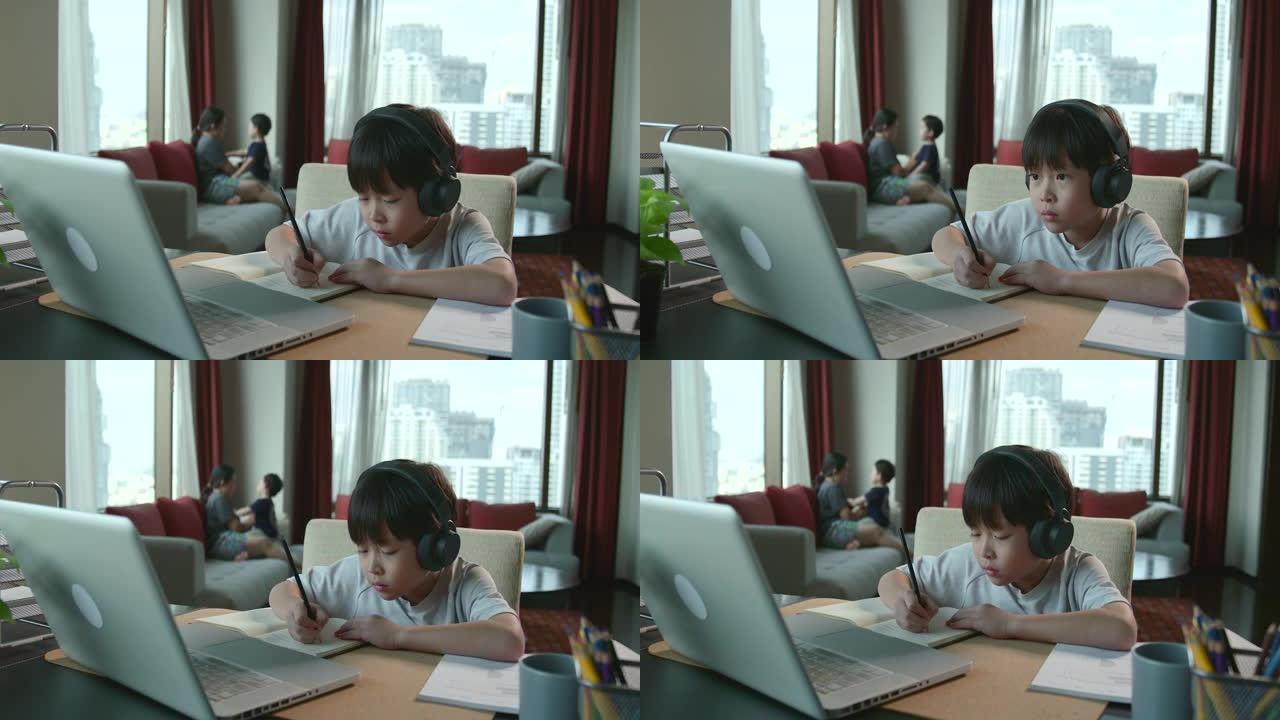 亚洲男孩正坐在家里专心在线学习，他记下了自己所学到的知识。通过笔记本电脑，人们因为covid 19而