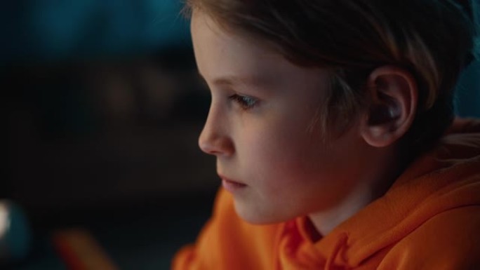 在家里一个黑暗舒适的房间里，一个可爱的年轻少年男孩被集中在电脑屏幕上的肖像。他穿着橙色连帽衫。好奇真