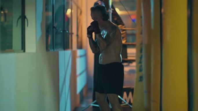 泰拳亚洲中国现役高级拳击手眼睛闭着练习拳击踢晚上在健身房的窗户旁边用耳机听音乐