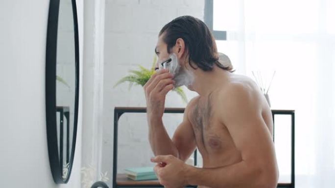 一个男人在浴室里看着镜子，用现代剃须刀刮胡须的慢镜头