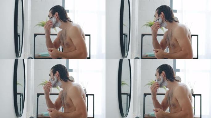 一个男人在浴室里看着镜子，用现代剃须刀刮胡须的慢镜头