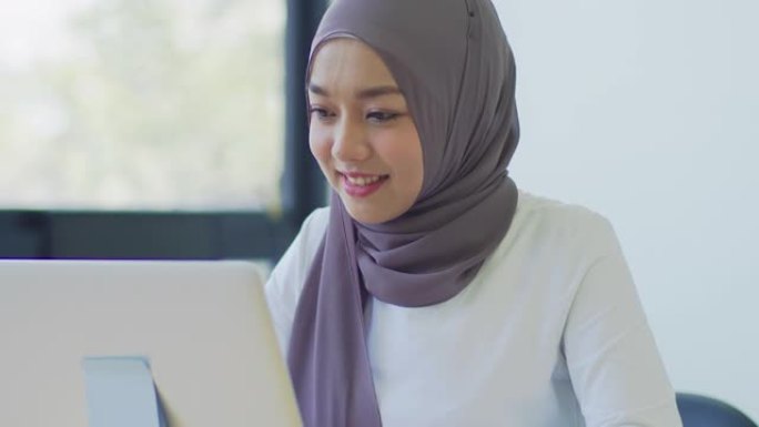 跟踪镜头: 带着头巾的快乐亚洲穆斯林妇女在家里或办公室工作和使用笔记本电脑，看着屏幕电脑，聊天，阅读