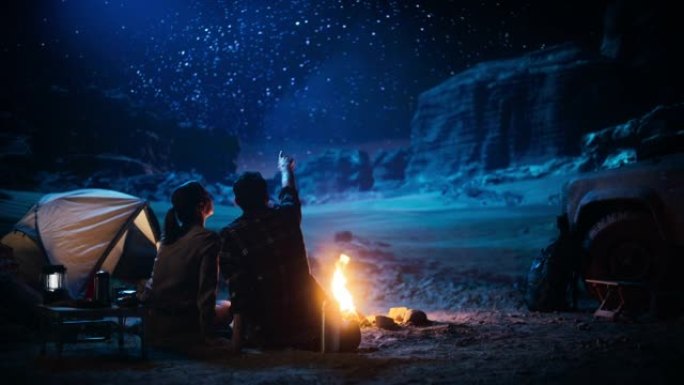 幸福的夫妇在峡谷露营，坐在一起看着篝火和星空，她把头靠在他的肩膀上。两个旅行的人在励志度假旅行中惊叹