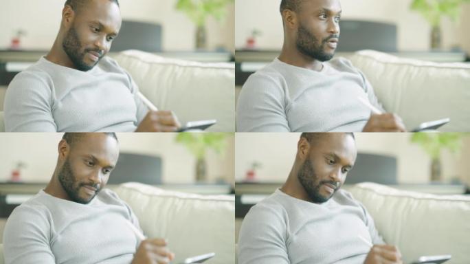 快乐的非洲民族年轻人在家用平板电脑工作，早上坐在沙发上放松。现代技术理念的人。
