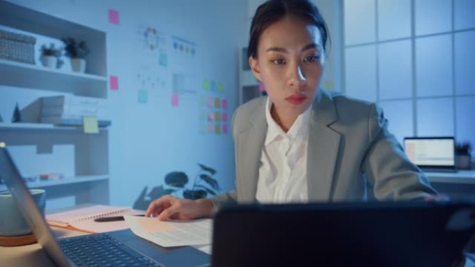 年轻的亚洲开朗的职业女性坐在办公桌上专注于笔记本电脑和数字平板电脑的工作，晚上在办公室检查详细工作。