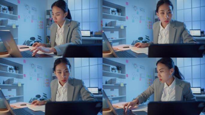 年轻的亚洲开朗的职业女性坐在办公桌上专注于笔记本电脑和数字平板电脑的工作，晚上在办公室检查详细工作。