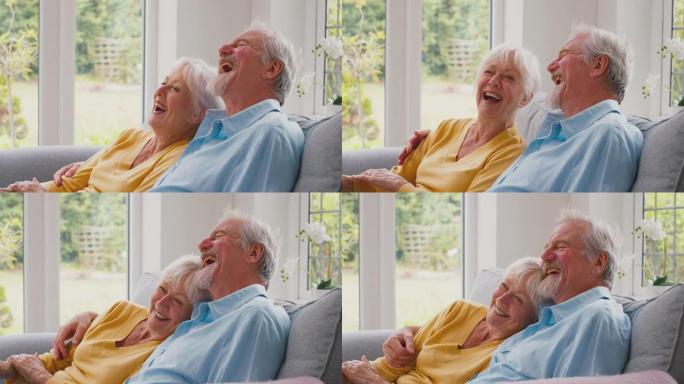 退休的高级夫妇坐在家里休息室的沙发上，一起观看摄像机电视和慢动作大笑