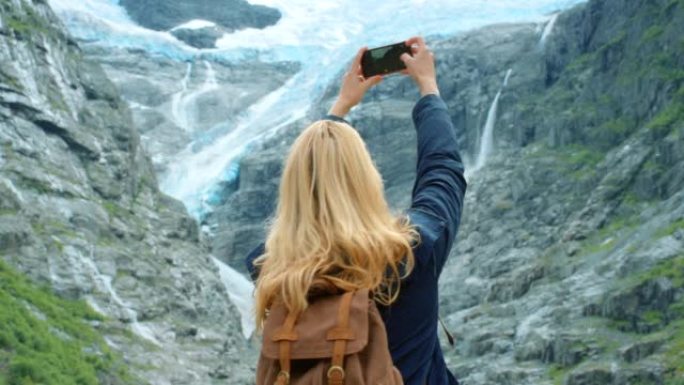 4k视频片段，一名年轻女子站着并在Briksdal Glacier的手机上拍照