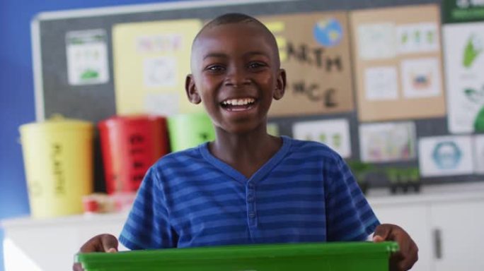 非洲裔美国小学生微笑着，拿着回收箱，站在教室里
