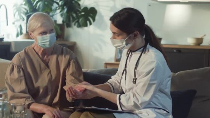 年轻的女医生穿着实验室外套检查70多岁的老年妇女在家里打电话拜访时戴着医用口罩的脉搏。