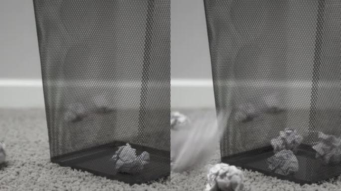 白色皱巴巴的纸球落在灰色背景上的灰色地毯地板上的金属废纸篮及其周围