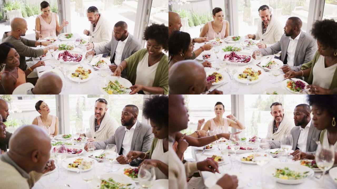 多样化和成熟的朋友群在家里的餐桌旁吃饭、喝酒和吃午餐。快乐的人在白天享受美食和美酒的同时微笑和大笑