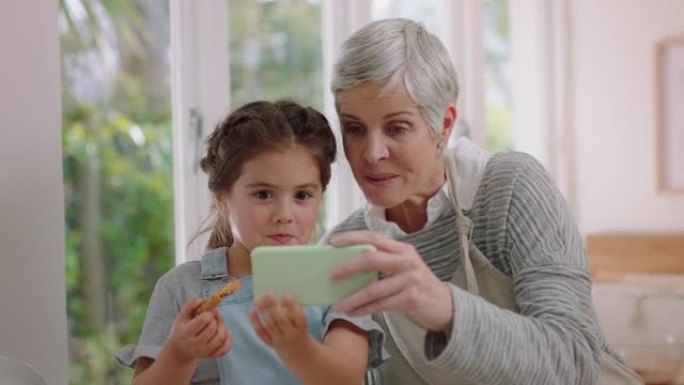 祖母和小女孩使用智能手机观看在线娱乐在厨房吃饼干快乐的孙女享受与奶奶分享周末在手机上浏览互联网