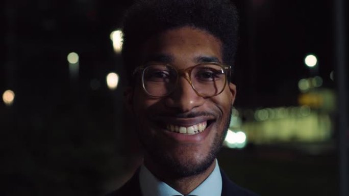 一个英俊时尚的黑人男性的特写镜头，戴着眼镜，晚上站在街上，看着相机，微笑着。穿着带有Bokeh灯光的