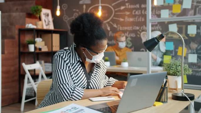 非裔美国女商人戴着医用口罩，使用笔记本电脑在办公室工作