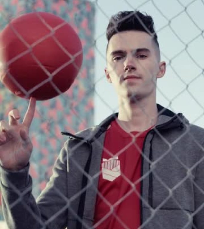 垂直屏幕: 时尚的年轻足球运动员摆姿势拍照，手指上旋转一个红球。英俊的足球运动员站在城市的篱笆后面，