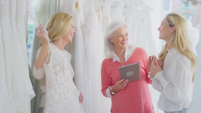 销售助理或商店老板用数字平板电脑帮助母亲和成年女儿在新娘商店选择婚纱-慢动作拍摄