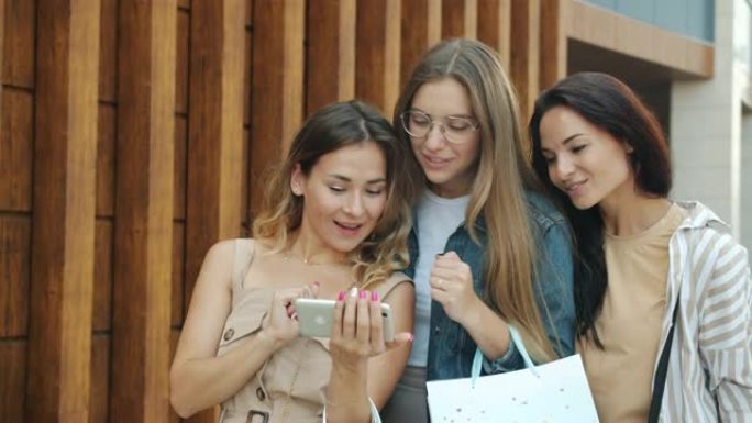 微笑的年轻女性朋友用纸袋摆姿势拍摄智能手机相机，然后在智能手机上观看照片
