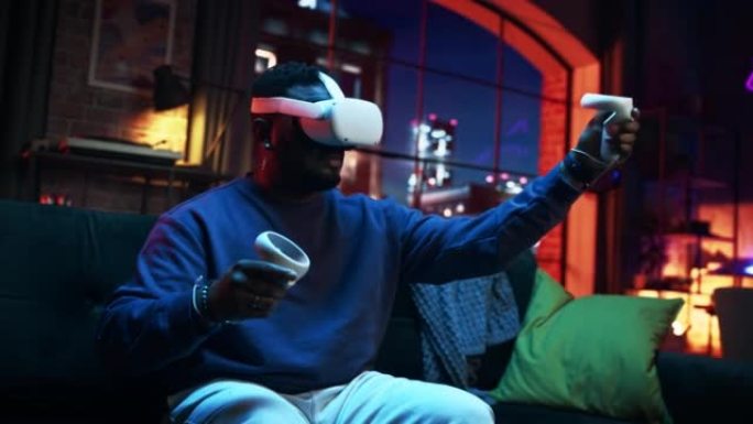 使用带有控制器的虚拟现实耳机拍摄的年轻时尚黑人的电影肖像。非裔美国男子晚上坐在家里的沙发上，玩VR电