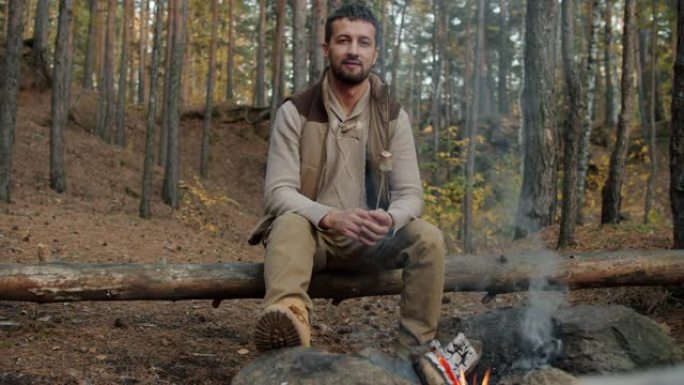 男性游客的肖像坐在树林里的火旁，拿着烤棉花糖微笑