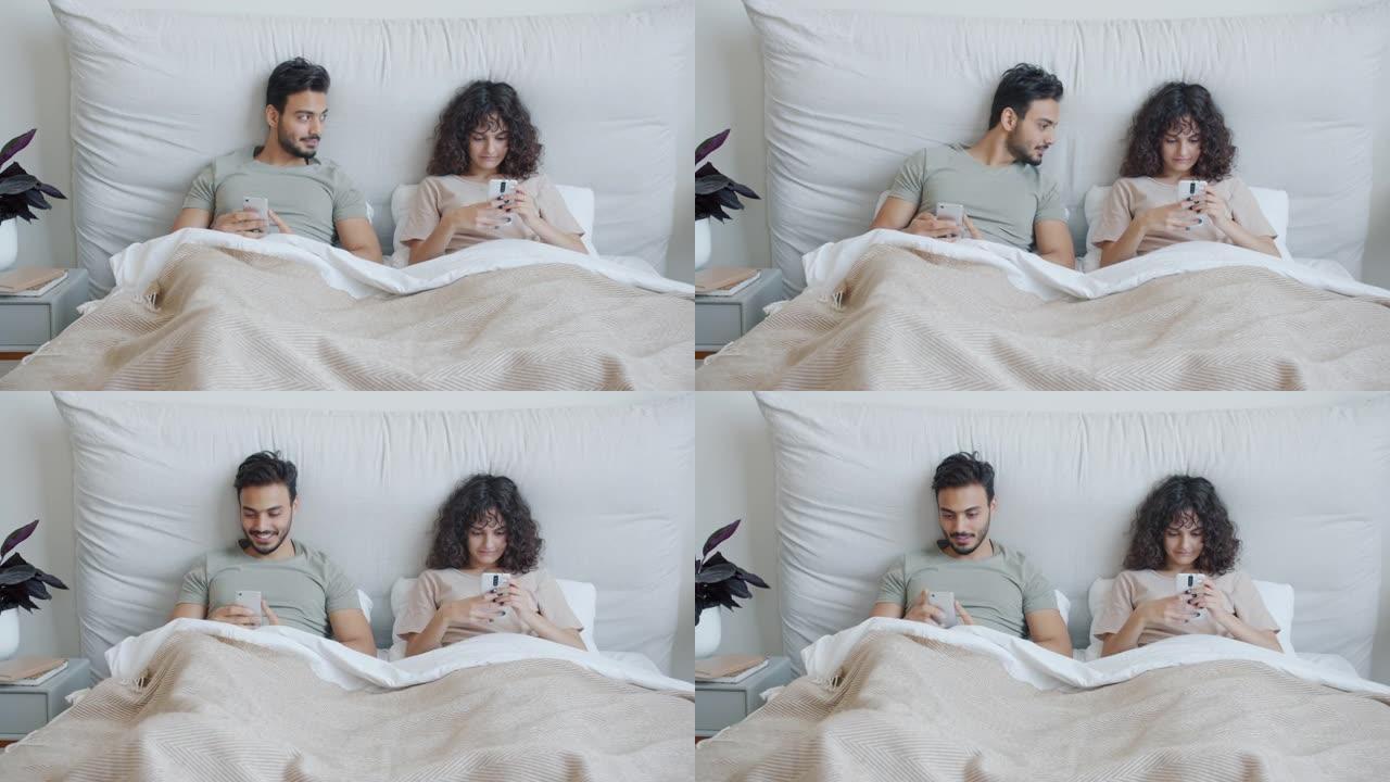 阿拉伯男人和高加索女孩一起躺在床上用智能手机在家休闲放松