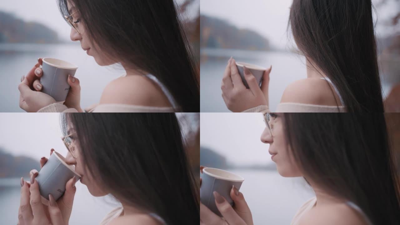 年轻女子从湖边的纸杯里喝热咖啡