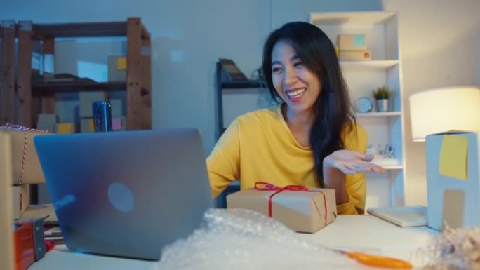 年轻的亚洲女商人使用智能手机接收采购订单，并在晚上在商店在线向客户视频直播显示产品包装。