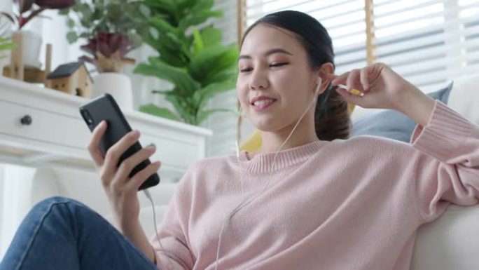 年轻的亚洲女人听播客在手机在线坐家里的沙发。