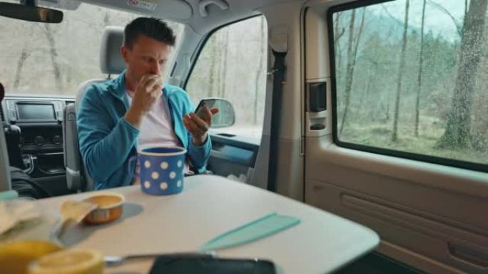 棕色短发的男人坐在面包车里，拿面包吃面包，放下面包，从早餐桌上拿一个咖啡杯和智能手机