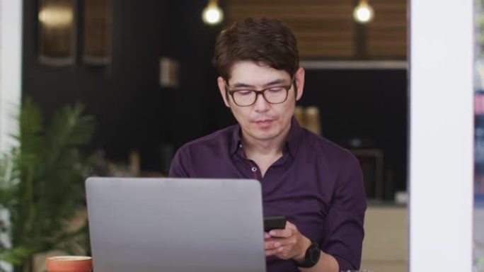 坐在咖啡馆里用笔记本电脑在智能手机上聊天的亚洲男子