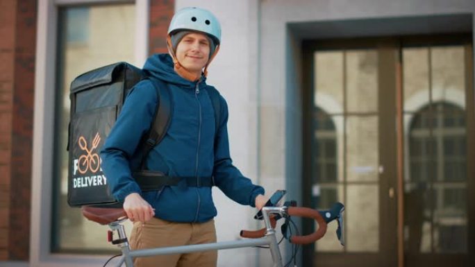 在时尚的现代市区，快乐的送餐男子戴着保暖背包和安全帽站在自行车旁边。微笑的快递员递送餐厅订单的肖像。