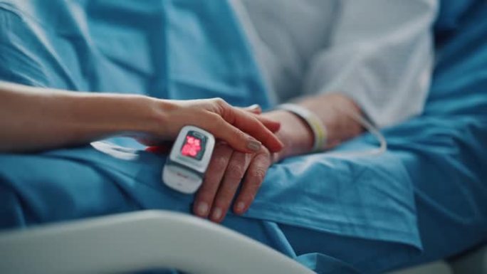 医院病房: 老年妇女用手指心率监测器躺在床上休息，有爱心的家人握着她脆弱的手，支撑和安慰。专注于手。