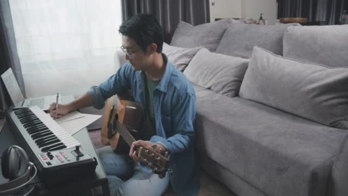 宽镜头亚洲成年男性在家里的客厅弹奏键盘钢琴和原声吉他。弹吉他创作音乐或在笔记本电脑上录制音乐的人。音