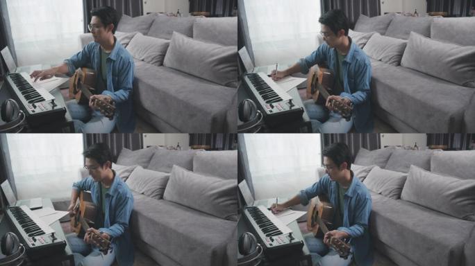 宽镜头亚洲成年男性在家里的客厅弹奏键盘钢琴和原声吉他。弹吉他创作音乐或在笔记本电脑上录制音乐的人。音