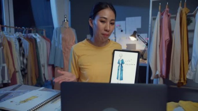 年轻的亚洲女士裁缝与平面设计师一起使用数字平板电脑会议，晚上在商店的桌子上讨论展示样品服装设计新系列