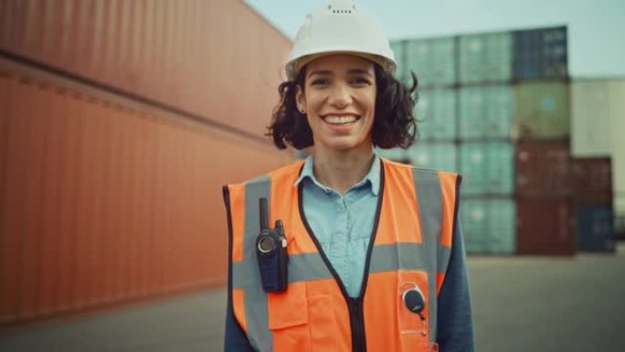 微笑的肖像，一位美丽的西班牙裔女工业工程师，穿着白色安全帽，安全背心，并在物流中心工作双向无线电。集